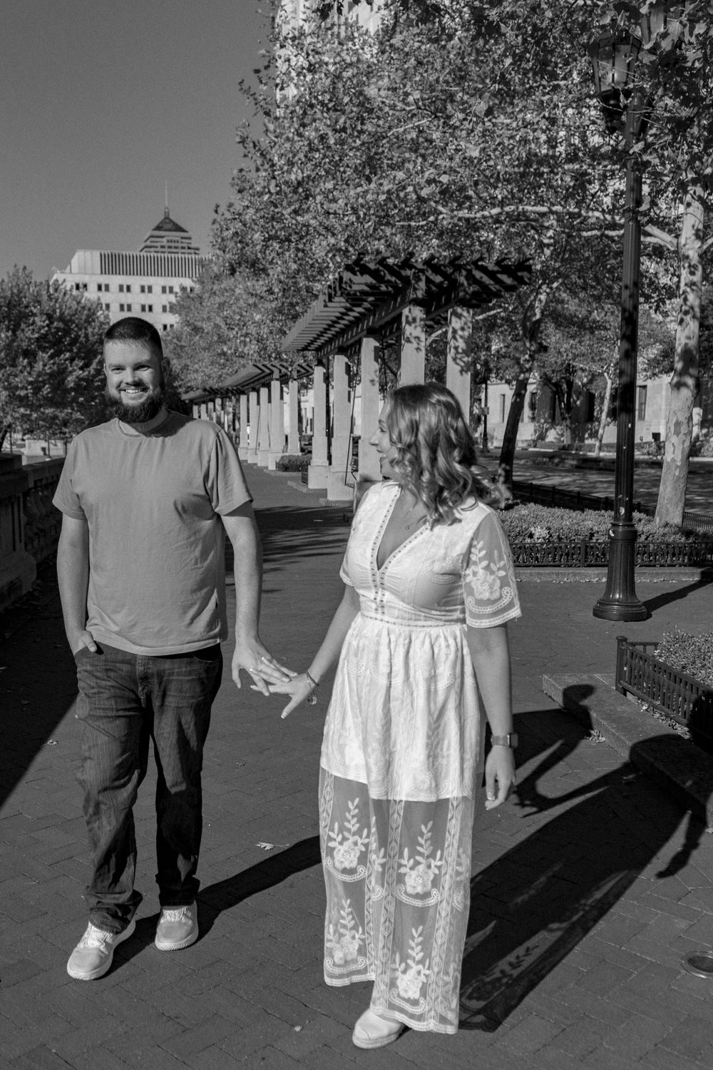 Two people standing together on a paved walkway with trees and buildings visible in the background.