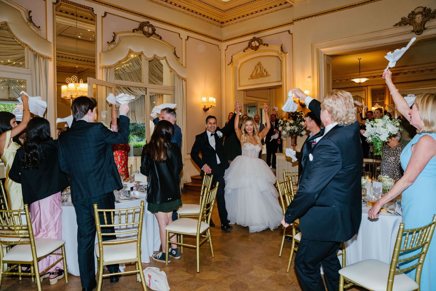 People dance and celebrate together during an elegant wedding reception in a grand ballroom with ornate gold details.