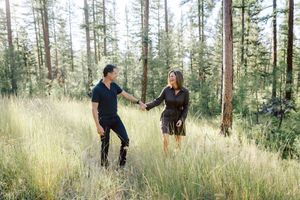 Two people playfully interact among tall grass in a pine forest setting.