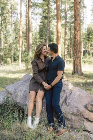 A couple sits together on a large rock formation in a pine forest.