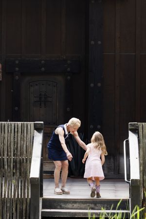 Elderly woman holding hands with a young girl on wooden steps outside a dark wooden building.