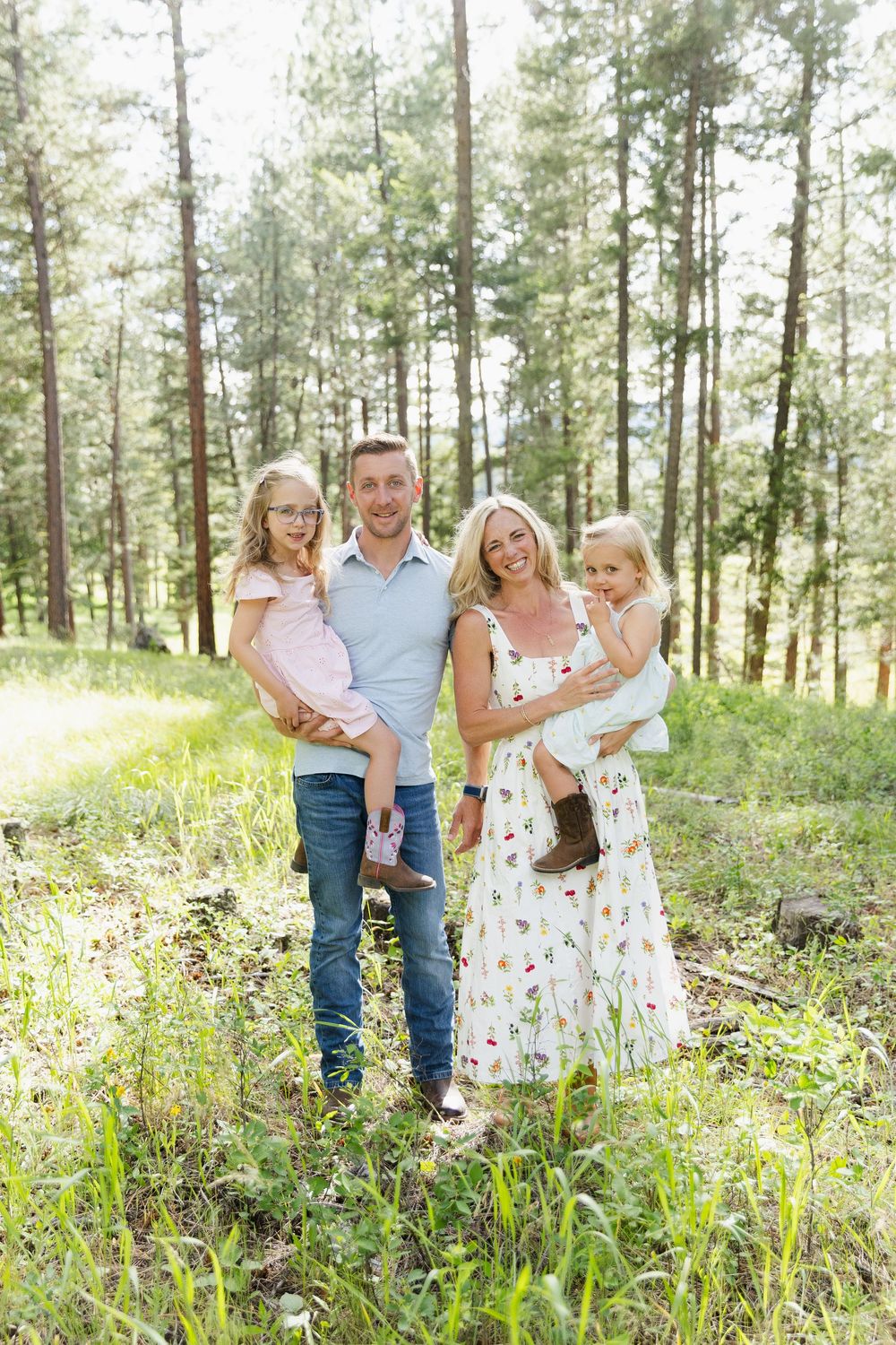 Happy family of four posing in a sunlit pine forest, parents holding two young daughters in casual summer outfits.