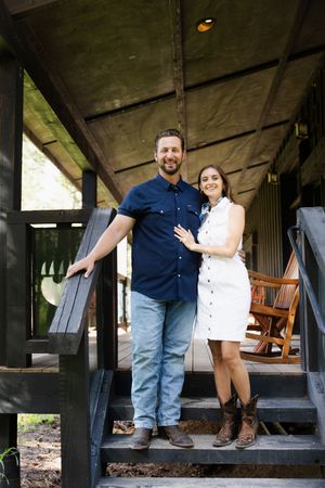 A smiling couple poses on wooden porch steps, man in navy shirt and jeans, woman in white dress and cowboy boots.