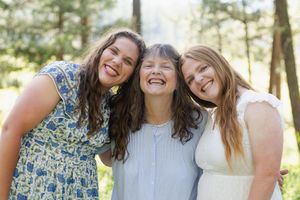 Three smiling women hugging outdoors in a sunlit forest setting, wearing summer dresses in a joyful group portrait.