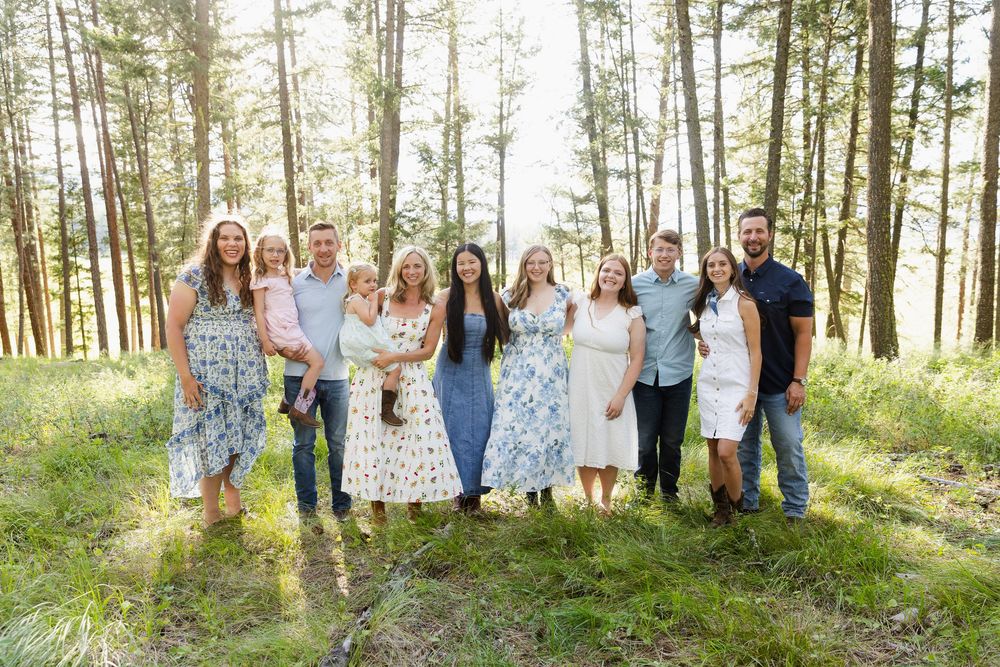 Large family group portrait outdoors in a pine forest, wearing coordinated outfits, smiling together on a sunny day.