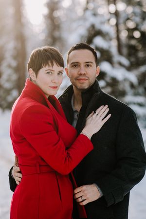 A couple embraces in a winter forest setting with one wearing a bright red coat.