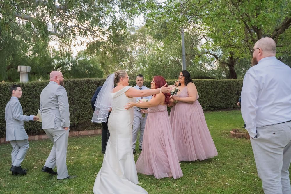 Wedding party members in pink dresses and grey suits dance together outdoors under trees during a celebration.