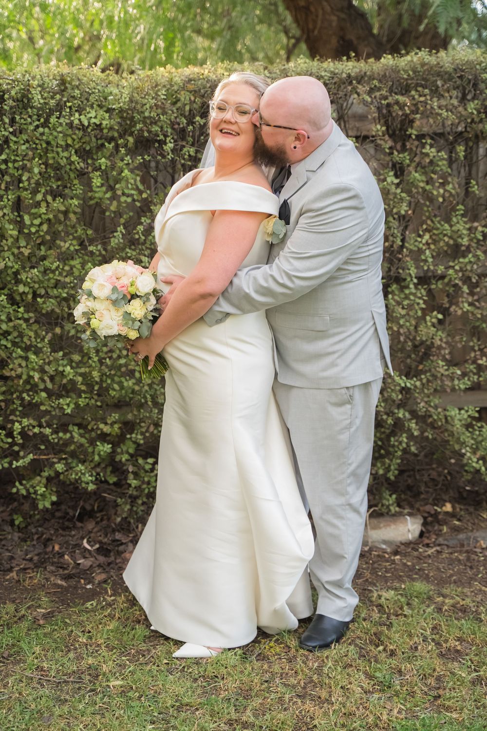 A romantic outdoor photo session of a couple embracing in front of lush greenery on their wedding day.
