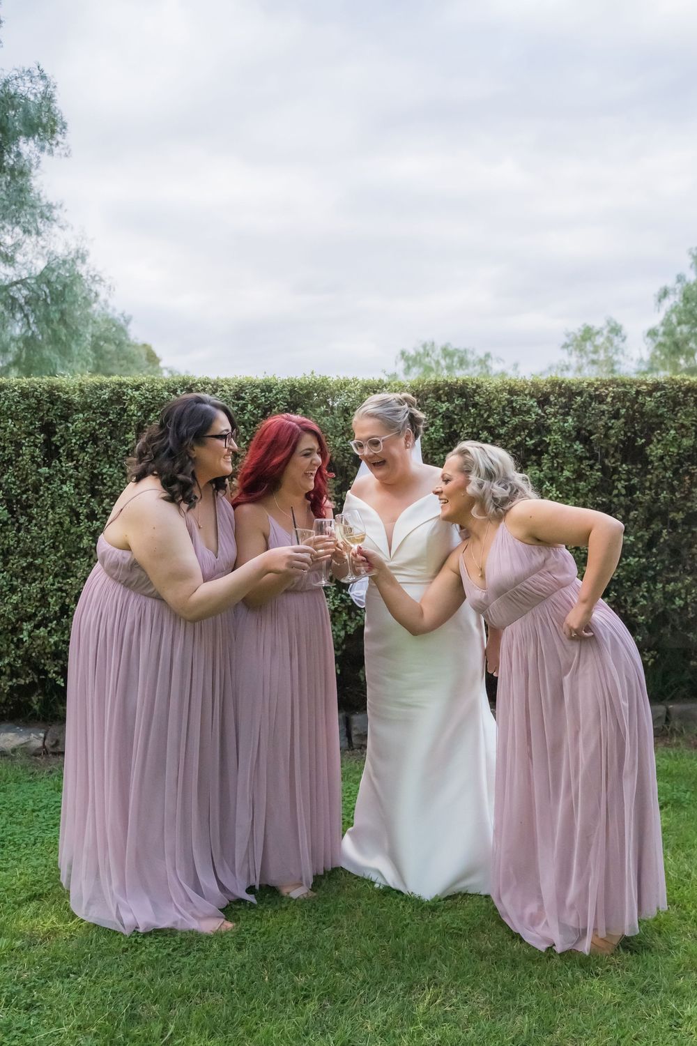 Wedding party in mauve bridesmaid dresses and white bridal gown pose playfully on grass.