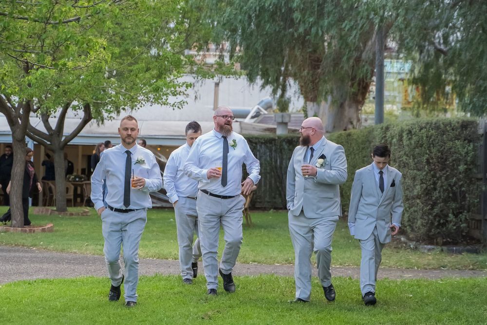 Groomsmen in light gray suits walk together across a grassy area outdoors during a wedding celebration.