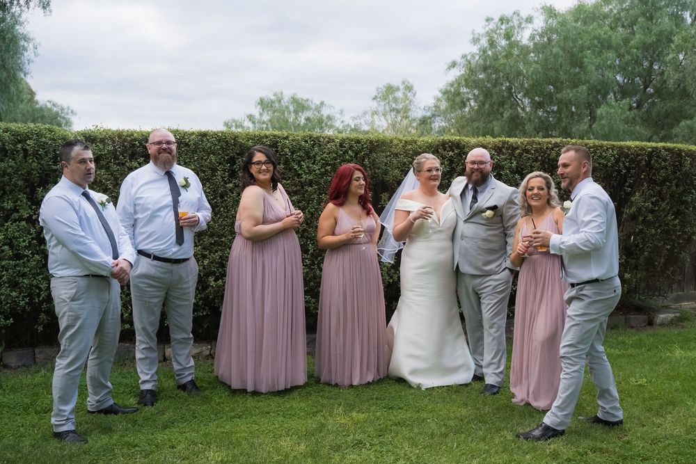 Wedding party members in light gray suits and pink dresses pose playfully in a garden setting with trees in background.