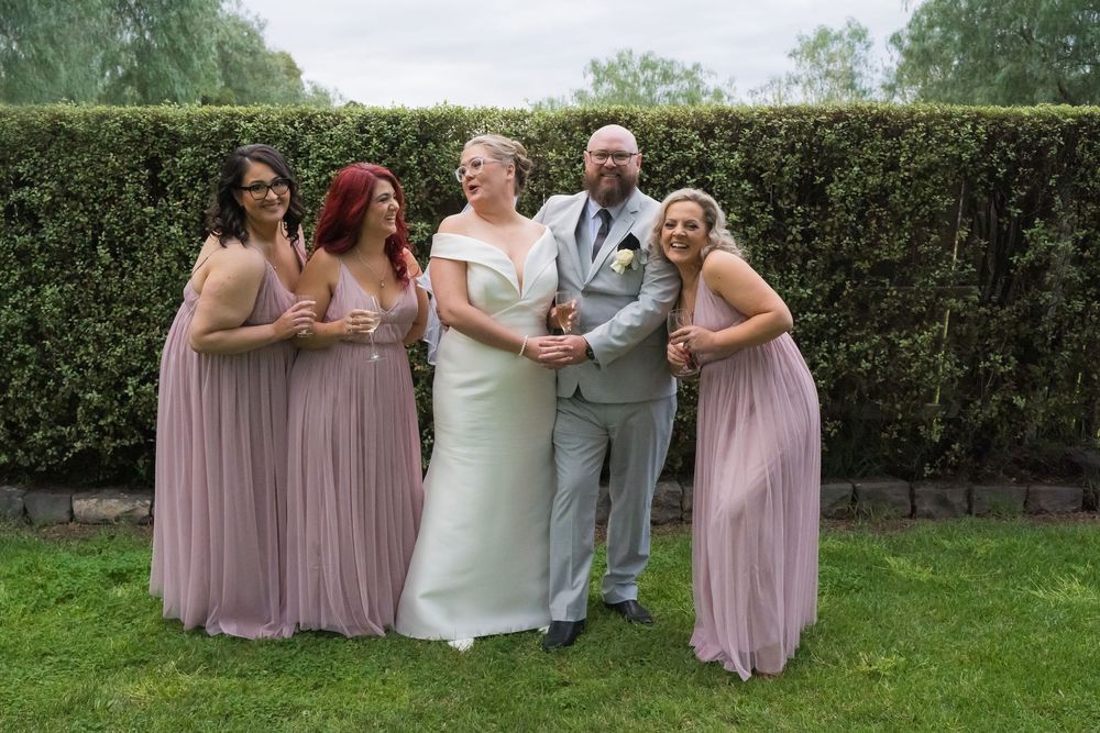Wedding party in pink dresses laughs joyfully together on a green lawn surrounded by hedges.