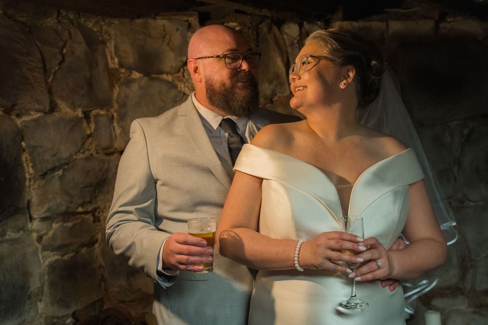 A romantic wedding couple shares intimate moments against a rustic stone wall while holding glasses and embracing.