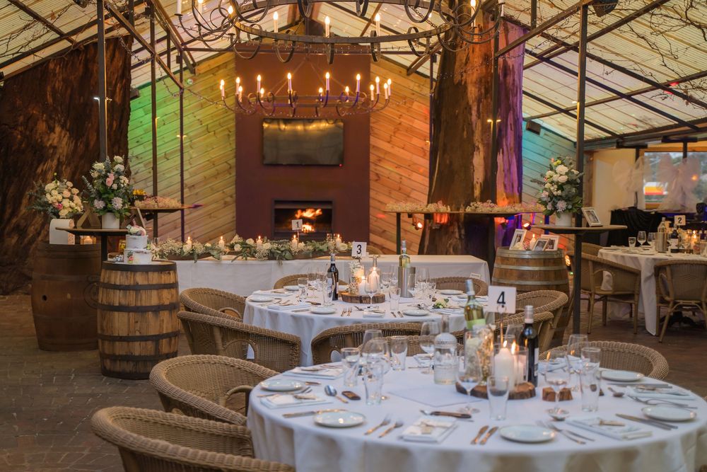 Elegant rustic event venue with round tables and chairs set up under wooden beams and sparkling chandeliers.