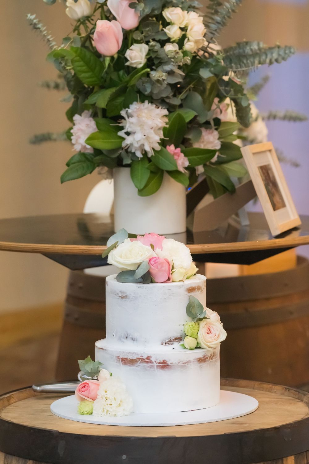Semi-naked wedding cake with fresh pink and white flowers topped with a matching floral arrangement centerpiece in white vase.