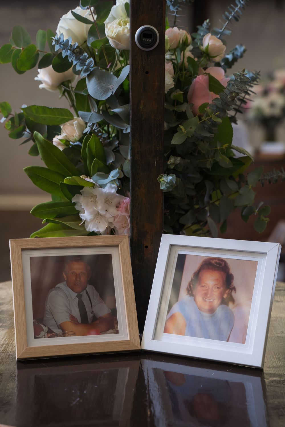Framed photographs displayed beneath floral arrangements with white flowers and green foliage in a dark setting.