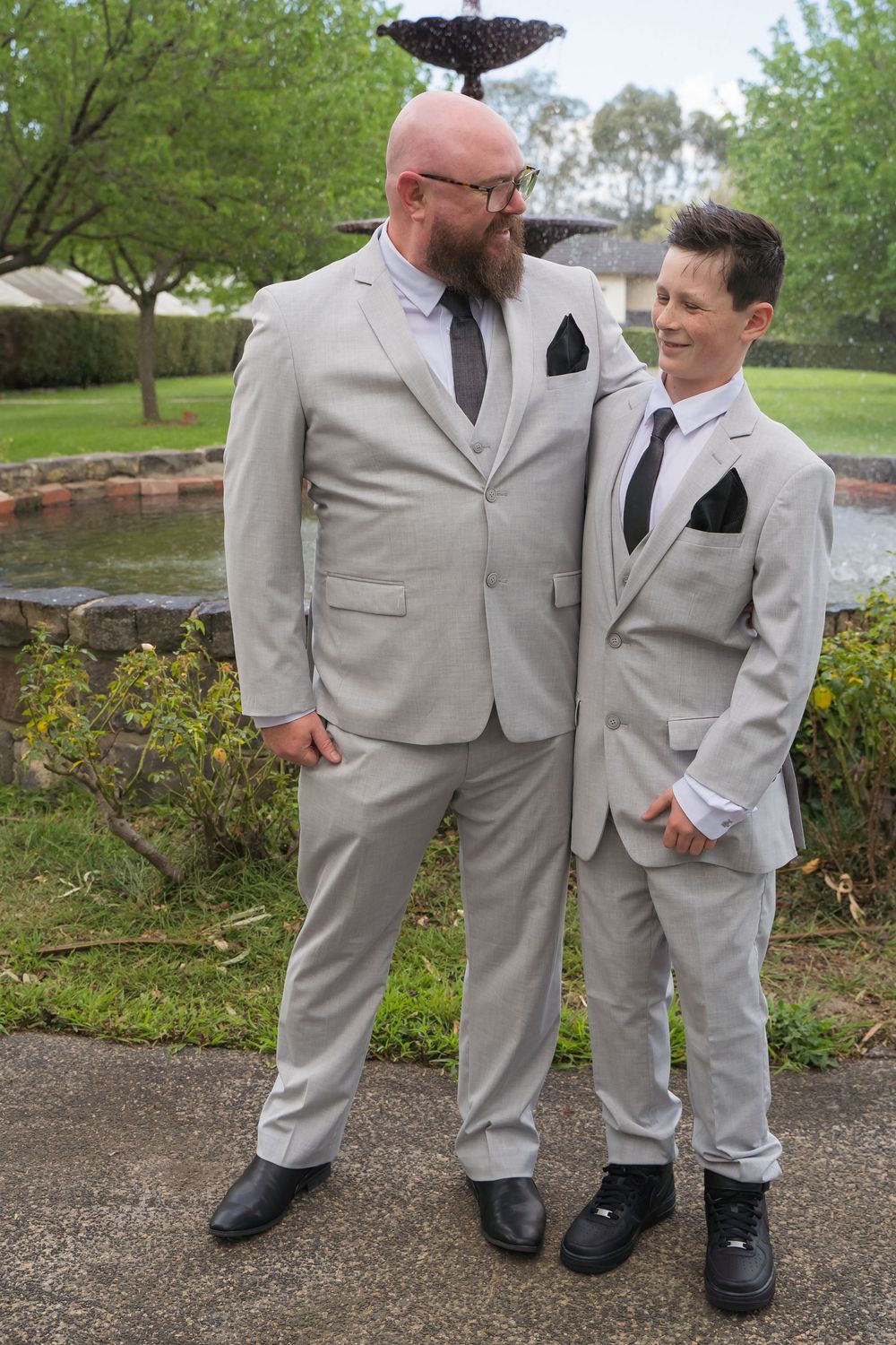 Two people in light gray suits share a lighthearted moment together outdoors while standing in a garden.
