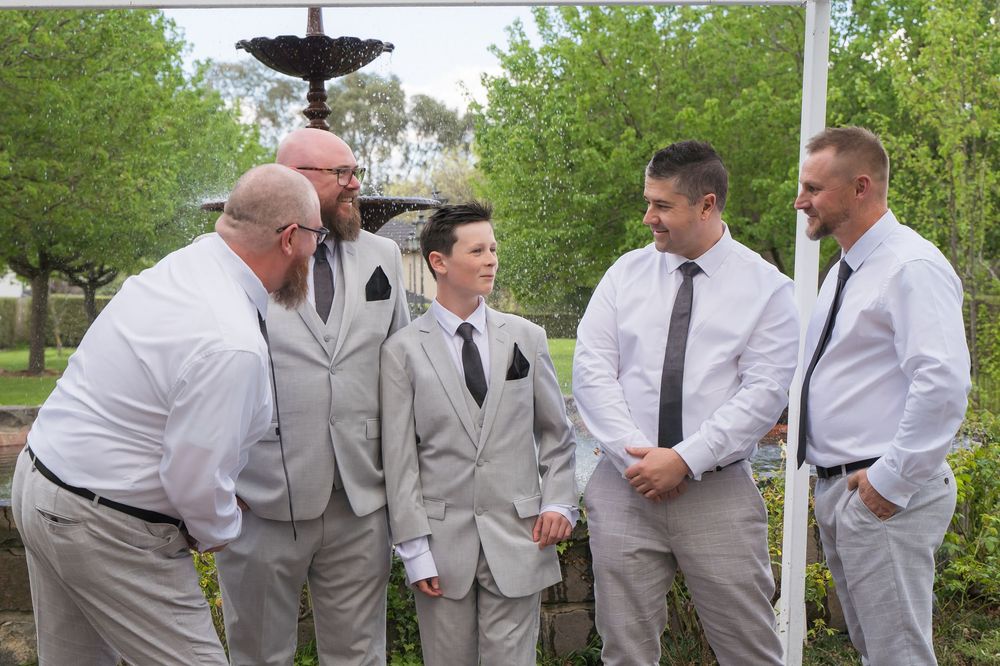A wedding party in light gray suits mingles outdoors near trees and bushes on a spring day.