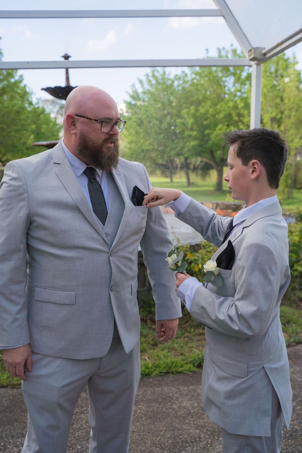 Groomsmen in matching light gray suits playfully adjust each other's boutonnieres during an outdoor wedding.