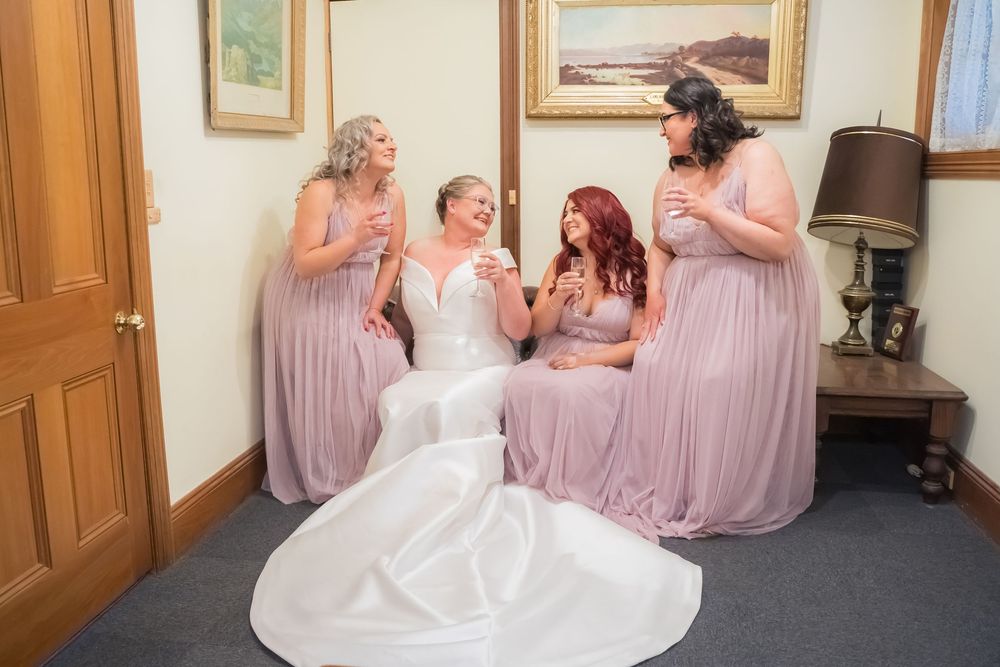 Bridesmaids in flowing blush pink dresses share a joyful moment with the bride in a white gown at a wedding venue.