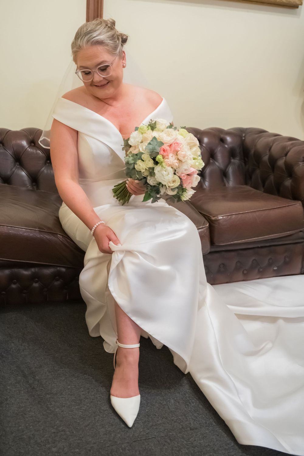 A bride in an elegant off-shoulder white gown sits on a brown leather couch holding a peach and white floral bouquet.