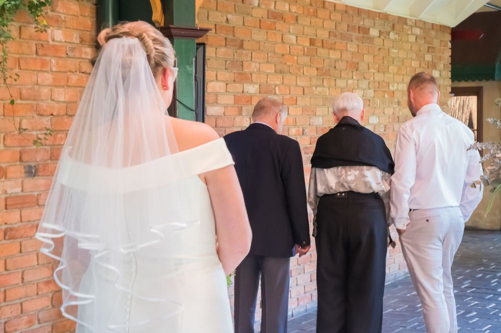 A line of people in white formal attire stand in front of a brick building with a visible decorative veil.