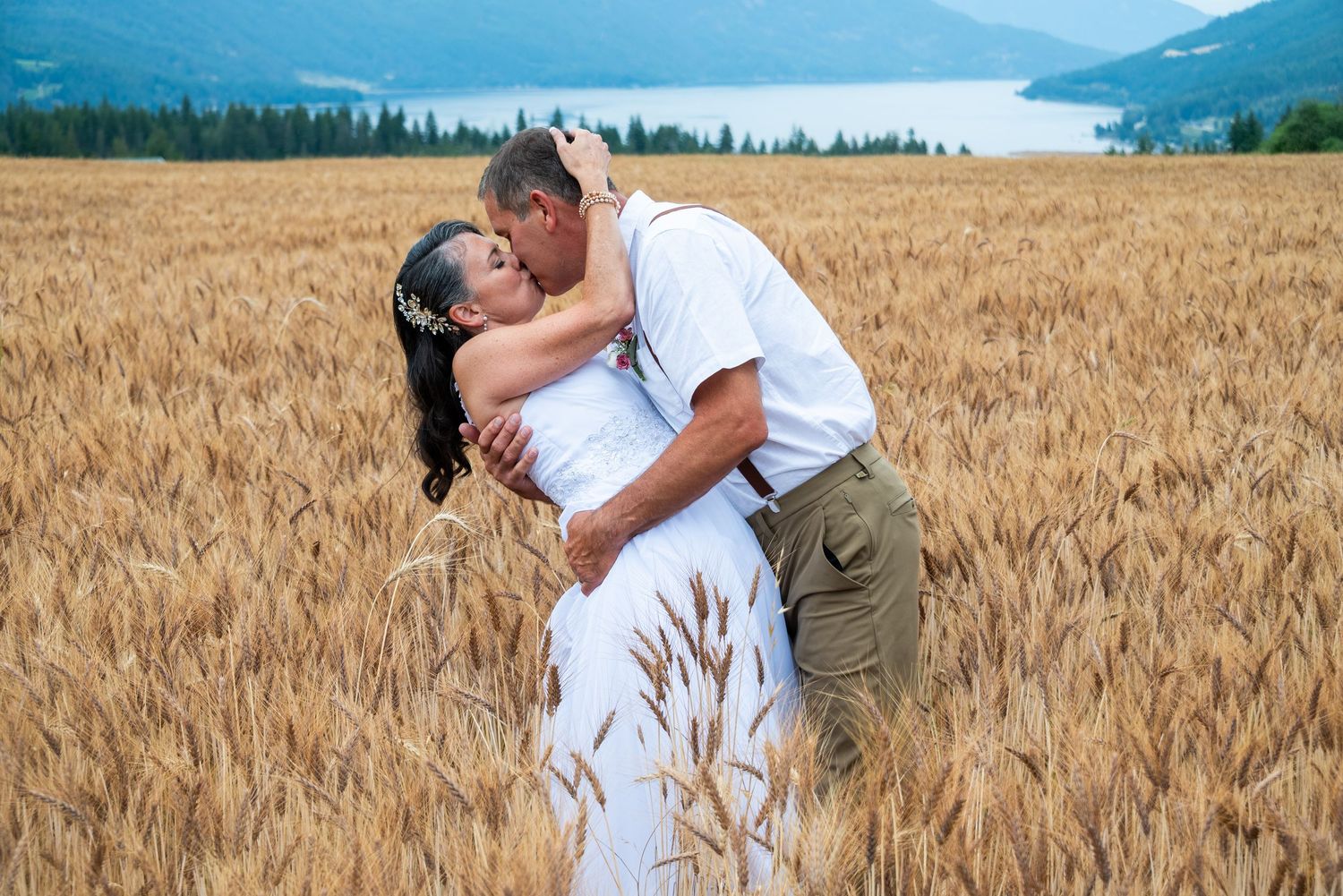 Wedding couple embracing in golden wheat field with mountain landscape background.