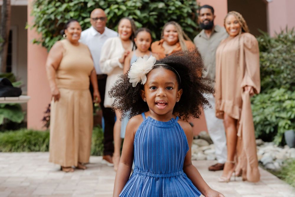 A young Black girl in a blue dress smiles in front of a family group wearing coordinated tan outfits outdoors.