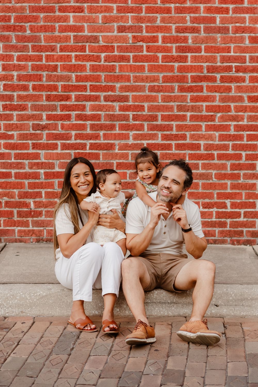 Happy family of four posing in front of a red brick wall, parents holding two young children while sitting on cobblestone steps.