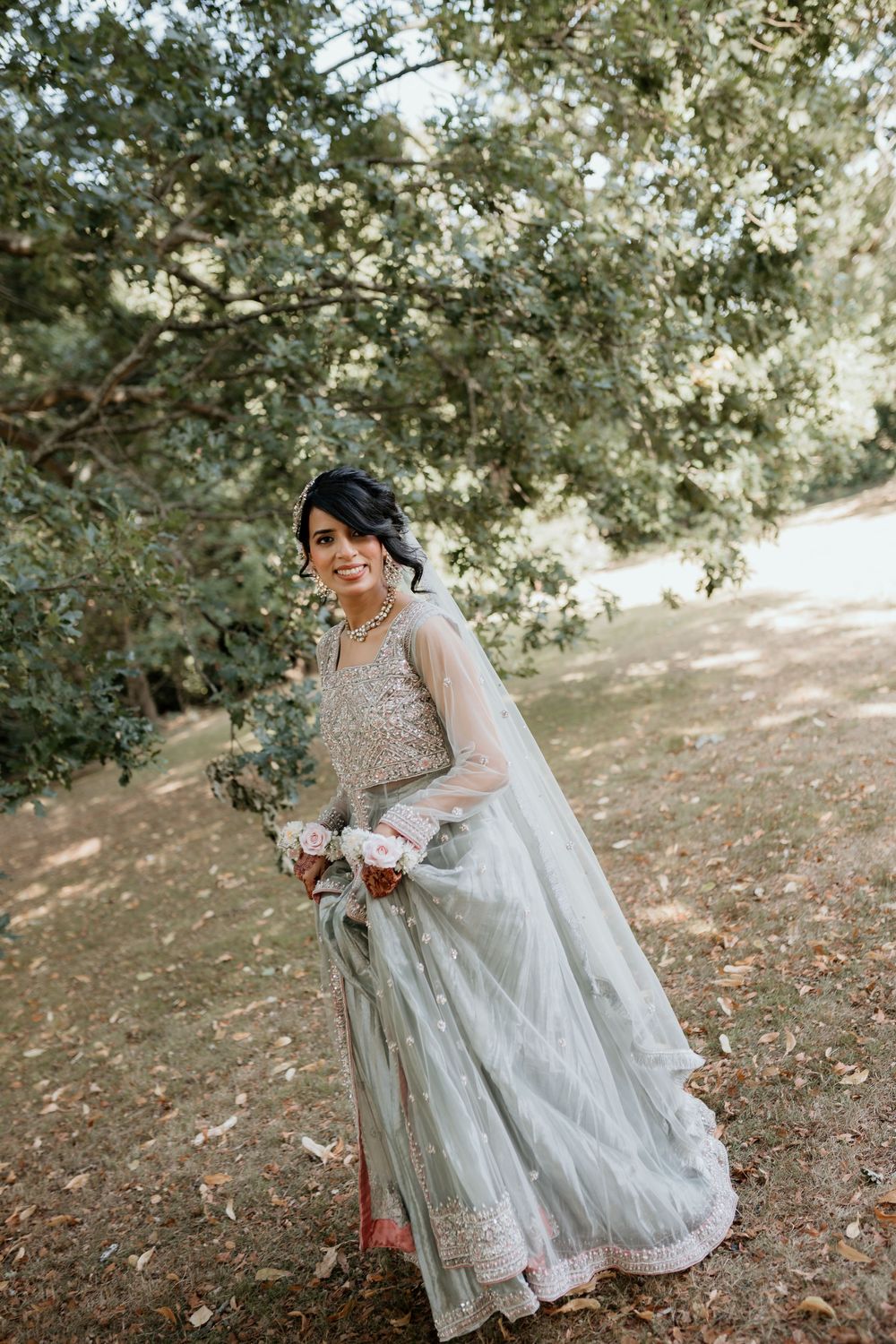 A person in a flowing white vintage-style dress and veil poses against a natural outdoor backdrop with trees and dappled sunlight.