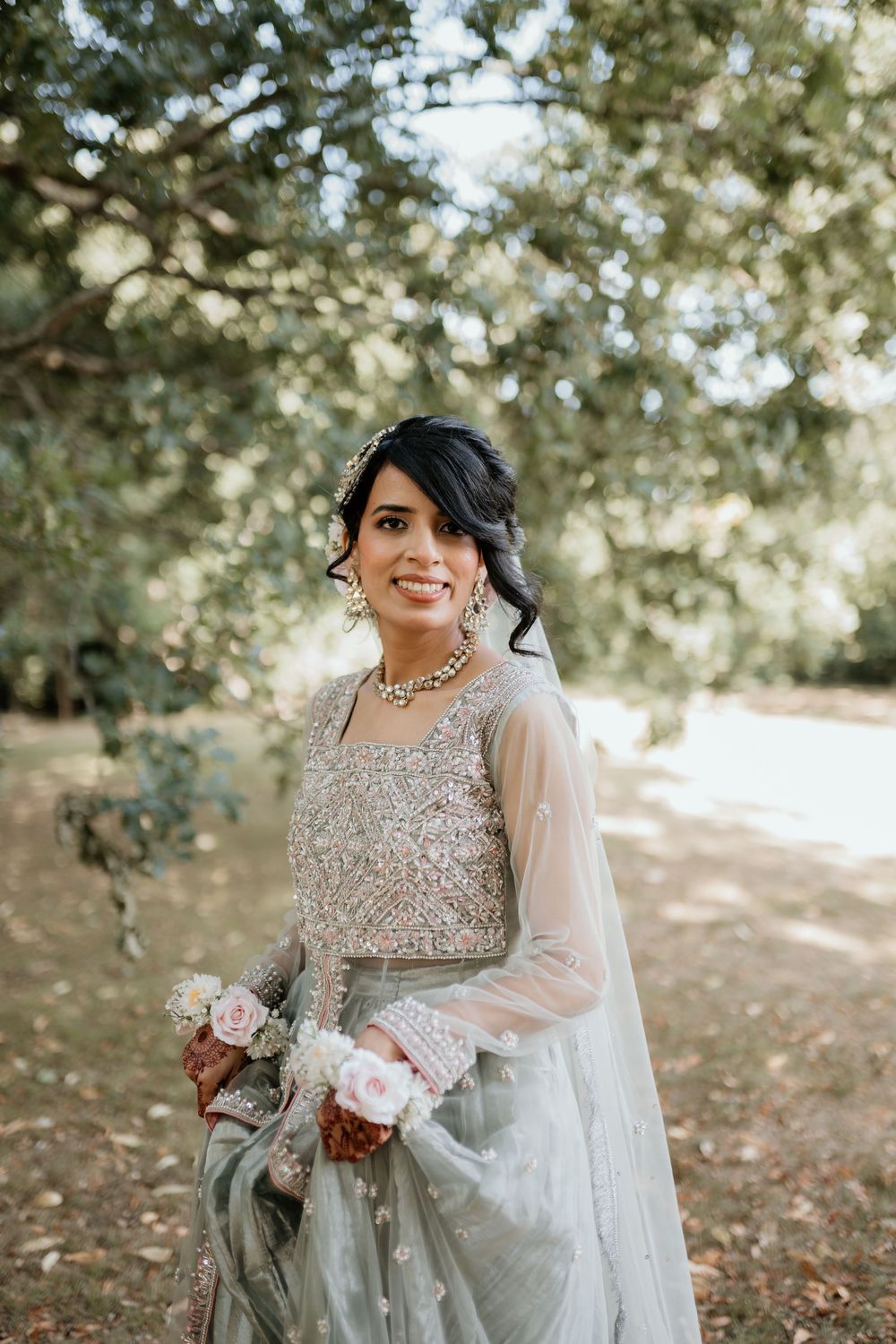 A bride in an elegant gray lace gown and veil poses outdoors among trees in natural lighting.