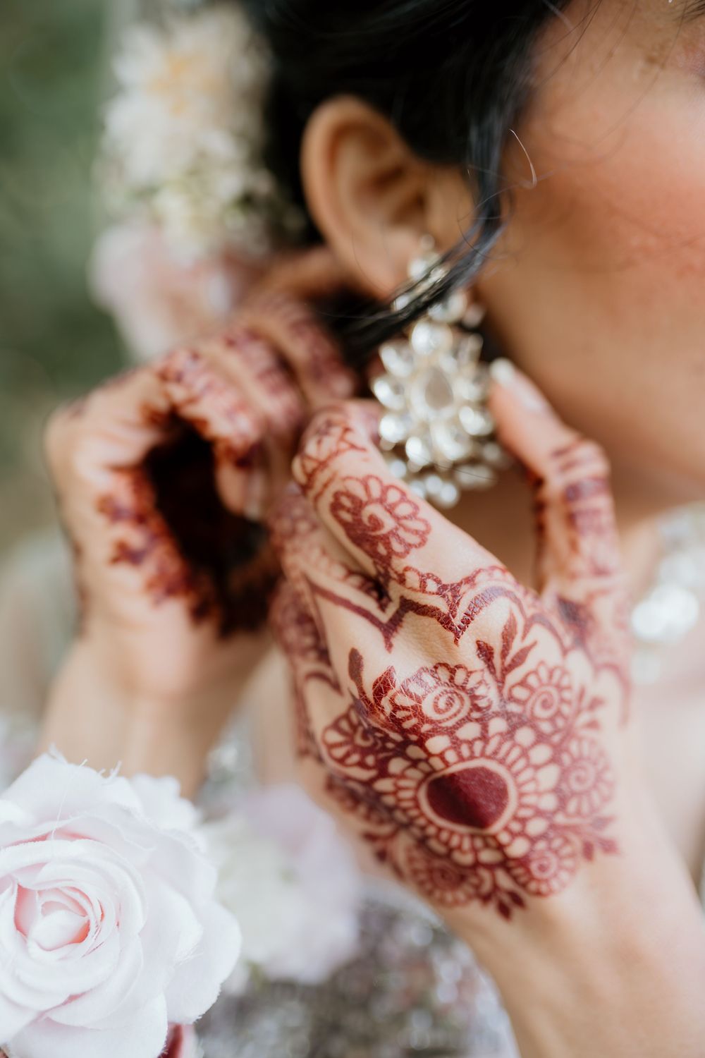 Beautiful henna design adorns hands holding a delicate pink rose while adjusting jewelry among white flowers.
