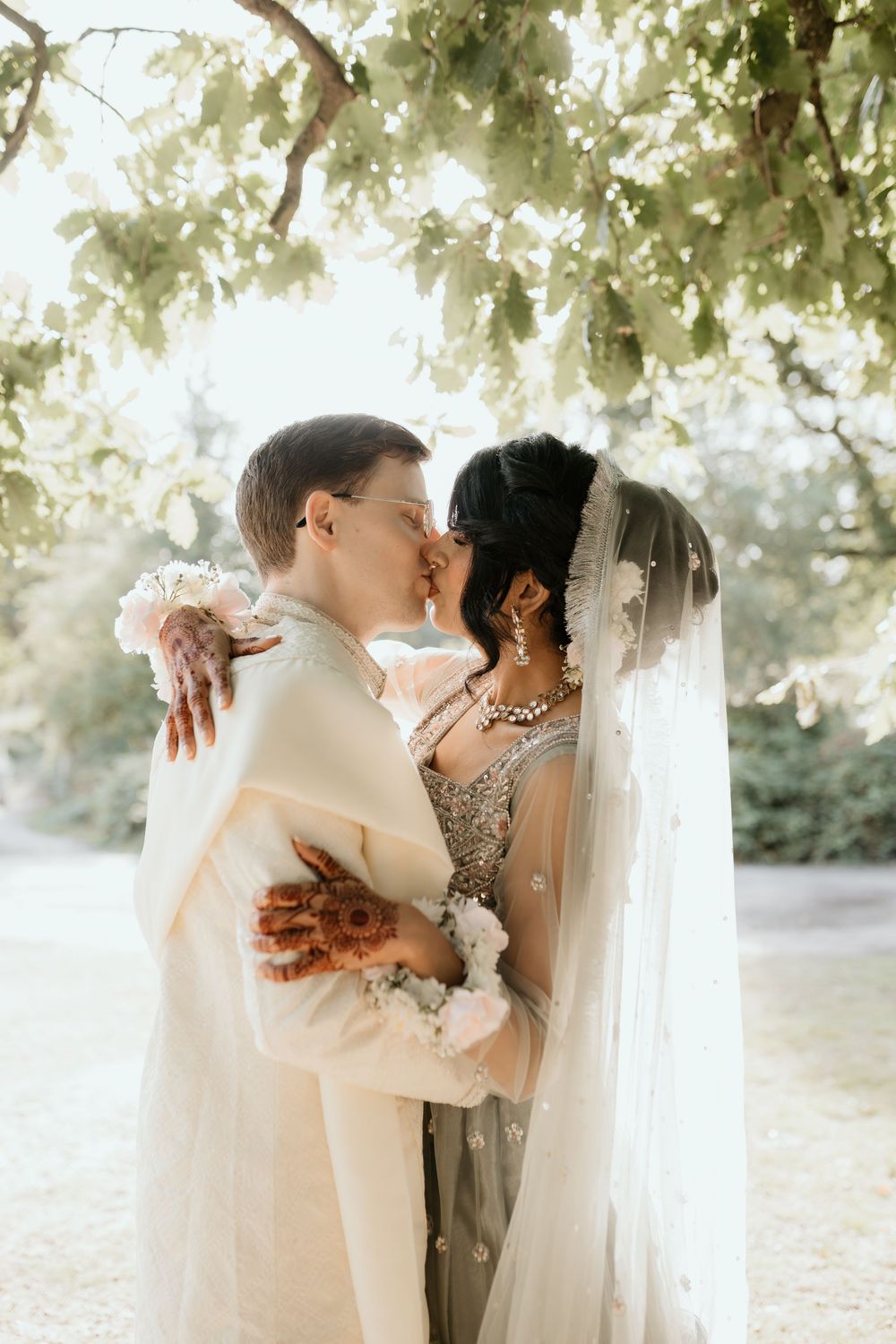 Wedding couple embraces tenderly against sunlit foliage backdrop wearing elegant attire.