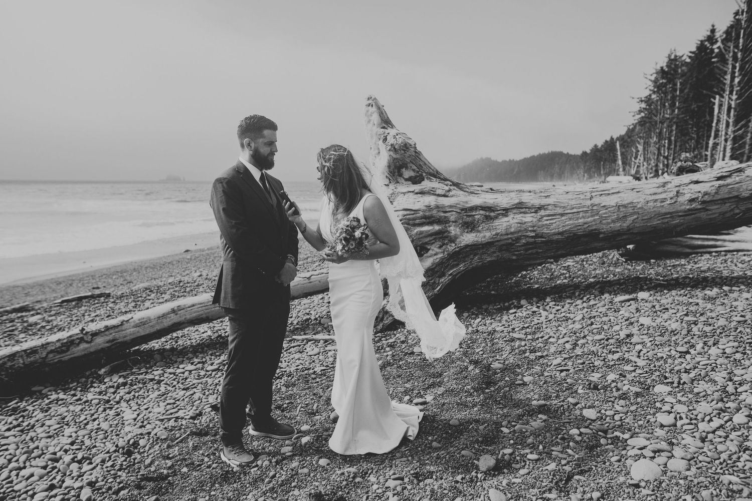 A couple in wedding attire shares a romantic moment on a driftwood-covered beach with evergreen forest in background.