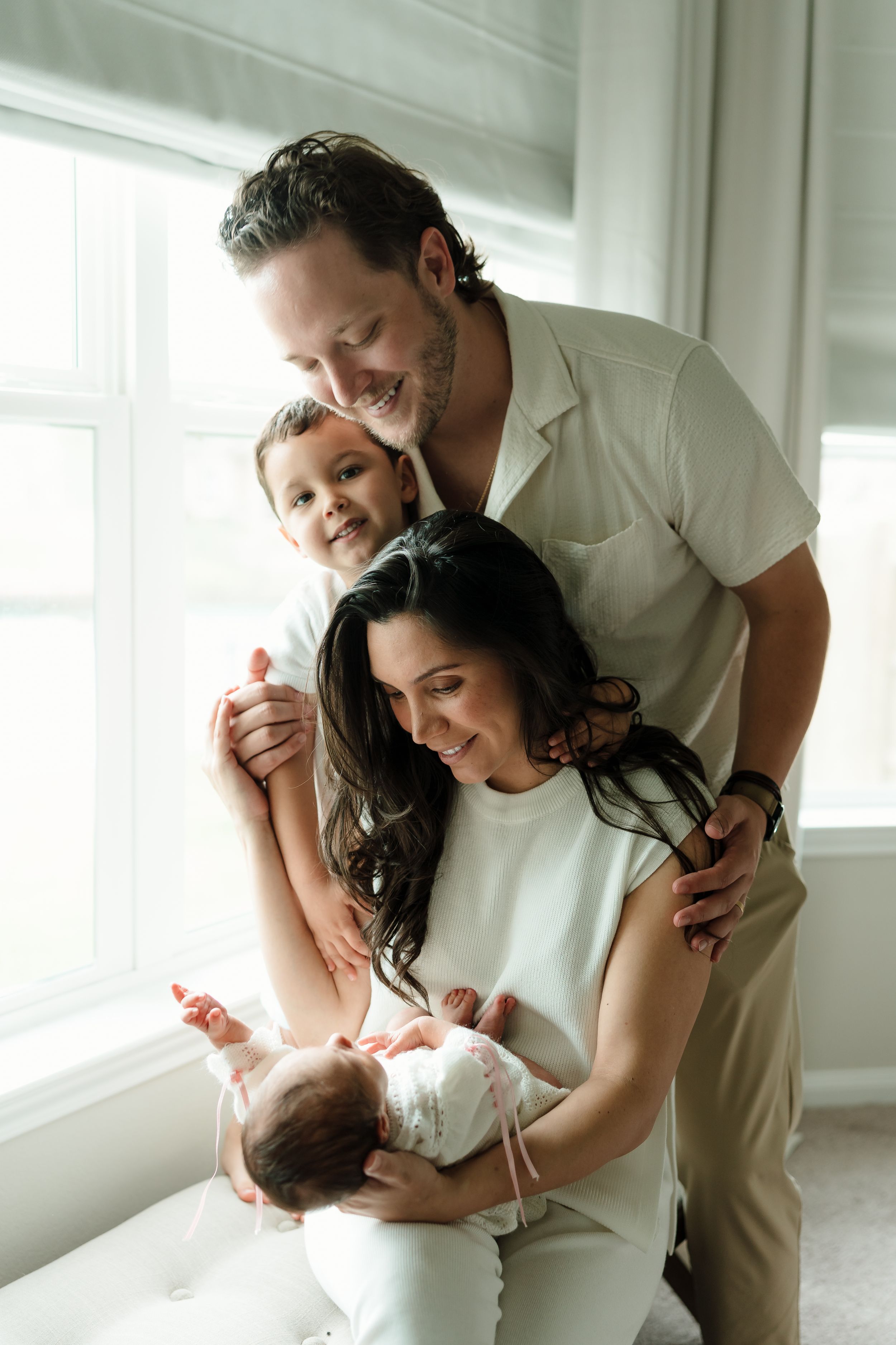 A happy family of four poses by a window, with parents smiling at their newborn baby while a young child joins the embrace.