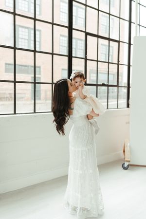 A woman in a white lace gown kisses a baby girl in white near large industrial windows.
