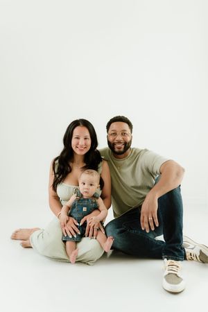 Happy interracial family portrait with mother, father, and baby in overalls sitting together on white background.
