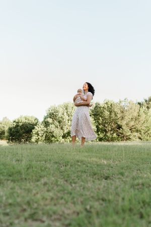 A woman in a floral dress holds her baby while standing barefoot on green grass in a park.