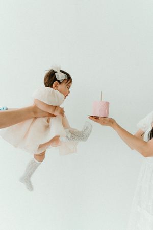 A baby in a pink dress reaches toward a pink birthday cake held by an adult, lifted in the air during a first birthday celebration.