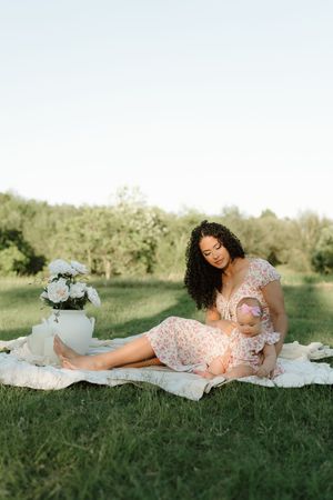 A mother in a floral dress sits on a blanket in a green park, cradling her baby girl near a white vase of flowers.