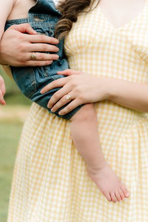Close-up of a woman in yellow gingham dress holding a baby in denim jacket, both wearing gold rings.