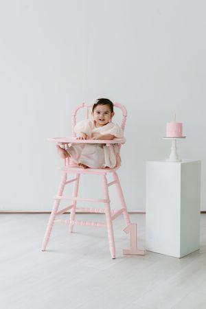 Baby girl sitting in pink high chair celebrating her 1st birthday with a pink smash cake on a white pedestal.