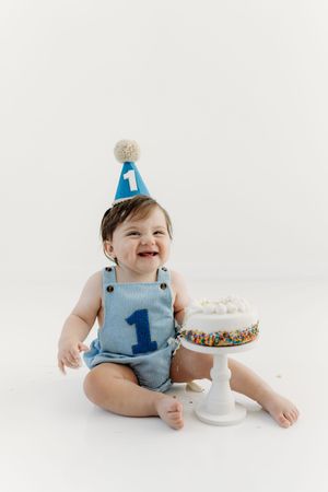 Baby boy in blue overalls and number 1 party hat smiling beside a sprinkle cake during first birthday smash.
