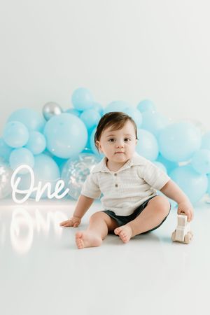 A baby boy sits on a white floor surrounded by blue balloons and a neon "One" sign for his first birthday.