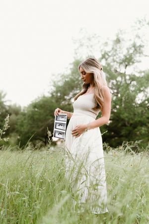 A woman in a white dress holds ultrasound photos in a grassy field.