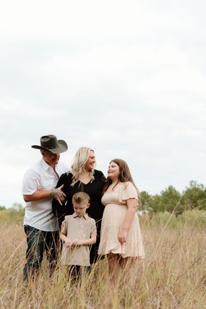 Family of four posing in tall grass field, father in cowboy hat, mother in black dress, two children smiling outdoors.