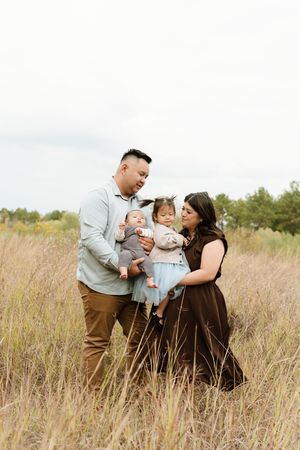 Family of four poses in tall golden grass field, parents holding baby and toddler girl in blue dress.