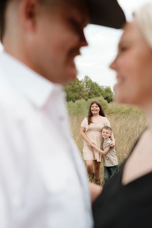 A family maternity photo with a pregnant woman and young boy in a field, framed by a blurred couple in the foreground.