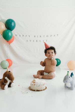 Baby Everett celebrates first birthday in party hat, smashing cake surrounded by colorful balloons and stuffed animals.