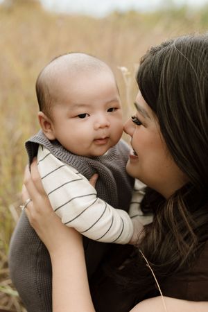 A mother tenderly kisses her baby outdoors in a golden field, holding the infant in a striped outfit and gray vest.